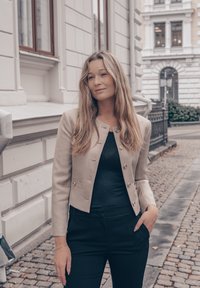 Blazer ajustado bege com botões dourados, usado sobre uma blusa preta. Calças pretas sob medida. O fundo apresenta calçamento de pedras e uma fachada de edifício.