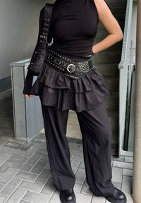 Μαύρο σύνολο που περιλαμβάνει τοπ ενός ώμου, φούστα με ρυτίδες και στρώσεις, φαρδύ μαύρο ζώνη με ασημένιες ανοξείδωτες λεπτομέρειες, και φαρδιά μαύρα παντελόνια με χοντρά μποτάκια.