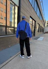 Μπλε ελαφρύ μπουφάν, μαύρο παντελόνι και άσπρα αθλητικά παπούτσια. Μαύρη τσάντα messenger κρεμασμένη στον ώμο. Αστική τοποθεσία με ανακλαστικά παράθυρα.