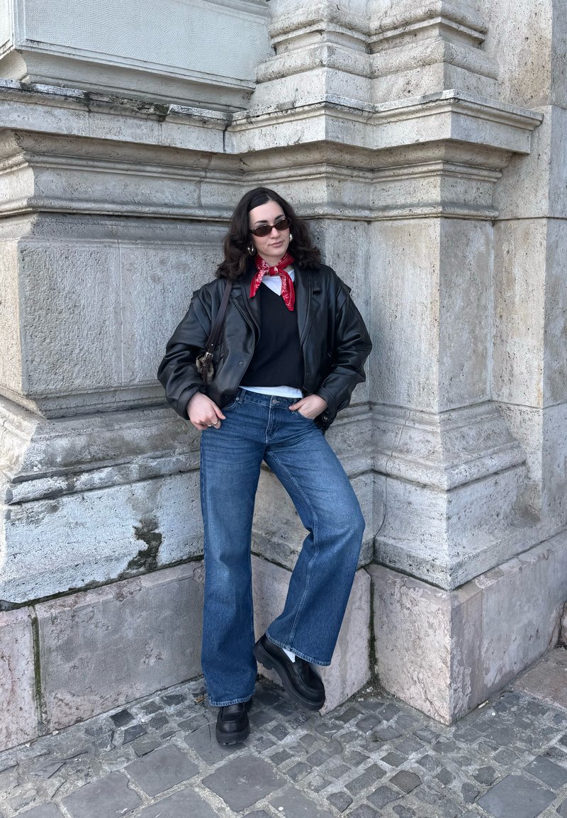 Donna con capelli ricci che indossa occhiali da sole, bandana rossa, giacca di pelle nera, jeans blu e stivali neri, in piedi contro l'angolo di un muro di pietra.