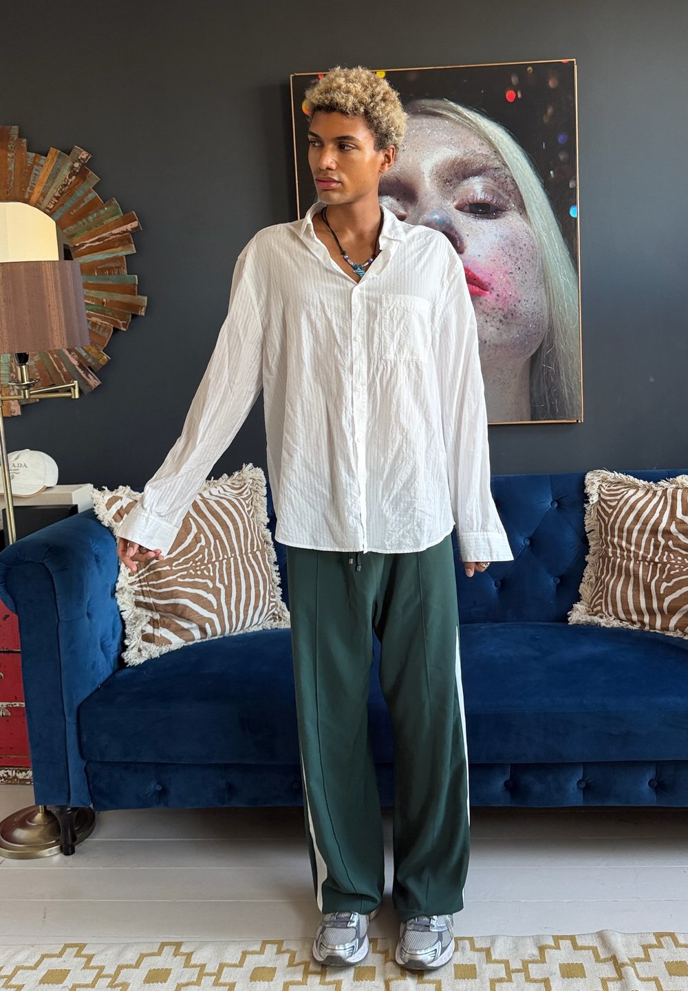 Personne aux cheveux blonds bouclés portant une chemise blanche et un pantalon vert se tient devant un canapé bleu avec des coussins à motifs et un grand portrait artistique.