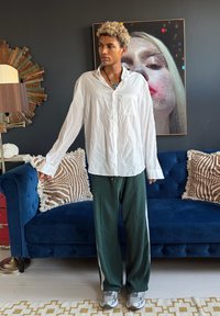 Personne aux cheveux blonds bouclés portant une chemise blanche et un pantalon vert se tient devant un canapé bleu avec des coussins à motifs et un grand portrait artistique.