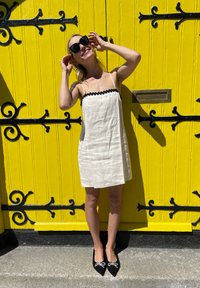 Witte linnen jurk met zwarte bies, zwarte puntige schoenen met strikaccenten, tegen een felgele houten deur met verfijnde zwarte hardware.