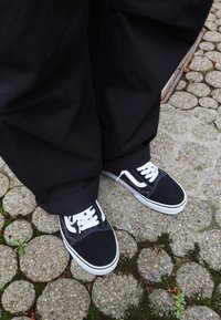 Osoba w czarnych spodniach i czarno-białych trampkach stoi na okrągłej kamiennej nawierzchni porośniętej mchem i małymi roślinami.