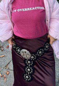 Maglietta rosa con testo bianco che recita "I ♥ OVERREACTING" abbinata a una lunga gonna di raso bordeaux scuro con una cintura decorativa nera con accenti in argento.
