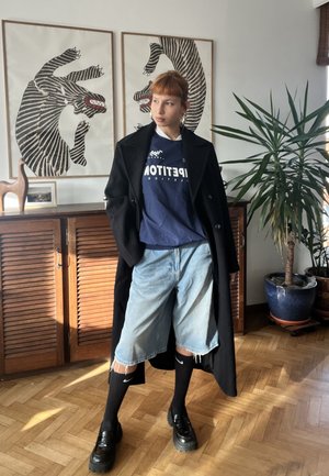 Persona joven con abrigo negro, camisa azul marino, shorts de mezclilla, calcetines negros Nike y zapatos de plataforma, de pie en una habitación con muebles de madera y planta.