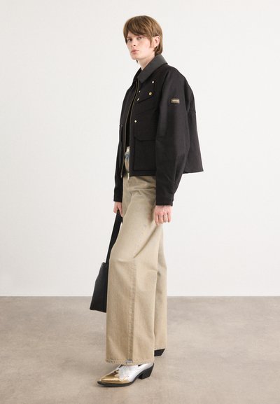 Chaqueta negra corta con herrajes dorados, pantalones anchos beige y zapatos metálicos. Diseño minimalista con acabado texturizado. Accesorio bolso negro.