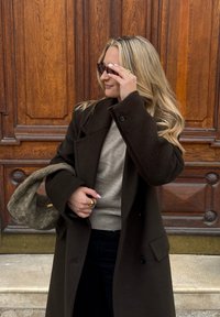 Długi, ciemnozielony płaszcz na beżowym swetrze, ciemne dżinsy i pleciona szara torebka. Ma na sobie ciemne okulary przeciwsłoneczne i złoty pierścionek.