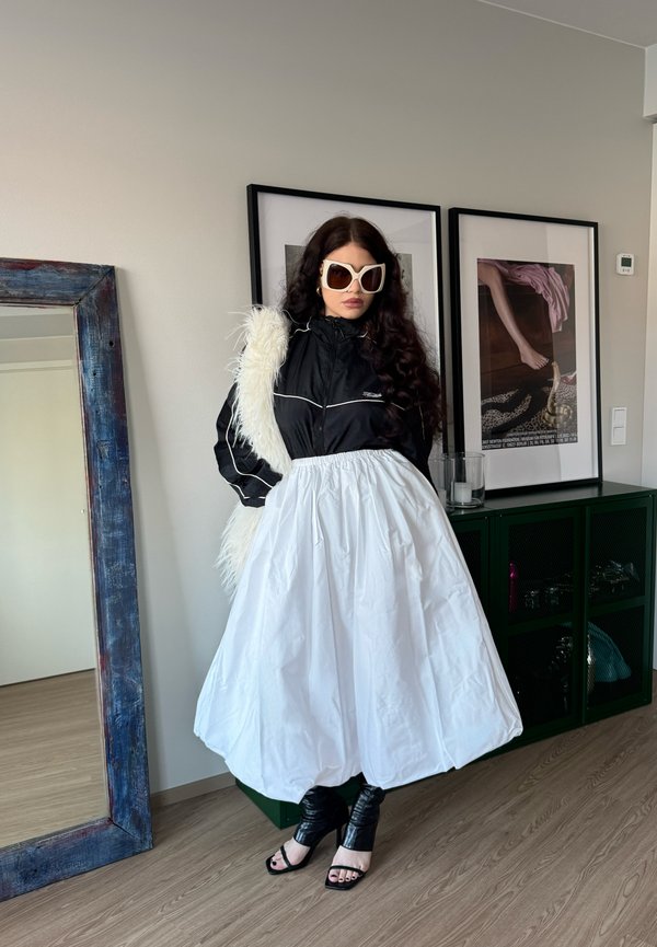 Kobieta w dużych białych okularach przeciwsłonecznych, czarnej kurtce, białej puchowej spódnicy i czarnych sandałach na wysokim obcasie stojąca obok lustra i oprawionej grafiki.