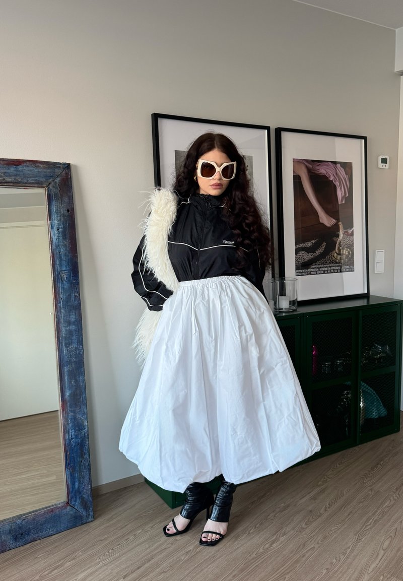 Kvinde iført store hvide solbriller, sort jakke, hvid puffet nederdel og sorte høje hæle, stående ved siden af et spejl og indrammet kunst.