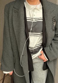 Osoba oblečená v vrstvenom sivom a čiernom kabáte cez pruhované tričko, s drôtovými slúchadlami a jednou rukou drží hnedú tašku.