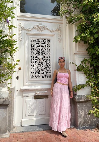 Falda maxi de lino rosa con diseño de volantes combinada con una camiseta ajustada rosa. Puerta decorativa blanca con intrincados detalles de hierro forjado y vegetación.