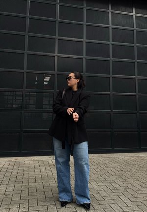 Mujer con gafas de sol ajustándose un blazer negro, lleva jeans anchos azules y zapatos negros puntiagudos, de pie sobre suelo pavimentado frente a una pared negra de rejilla.