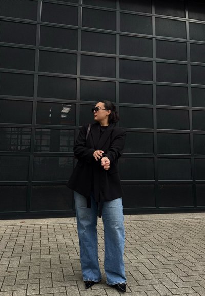 Mujer con gafas de sol ajustándose un blazer negro, lleva jeans anchos azules y zapatos negros puntiagudos, de pie sobre suelo pavimentado frente a una pared negra de rejilla.