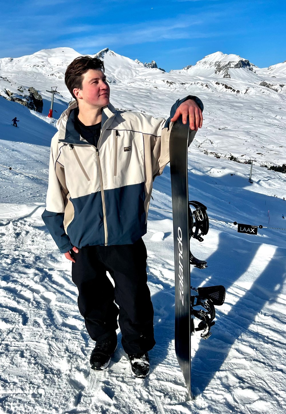 Hombre con chaqueta de invierno de pie en una pendiente nevada sosteniendo una tabla de snowboard en posición vertical, con montañas y telesillas al fondo.