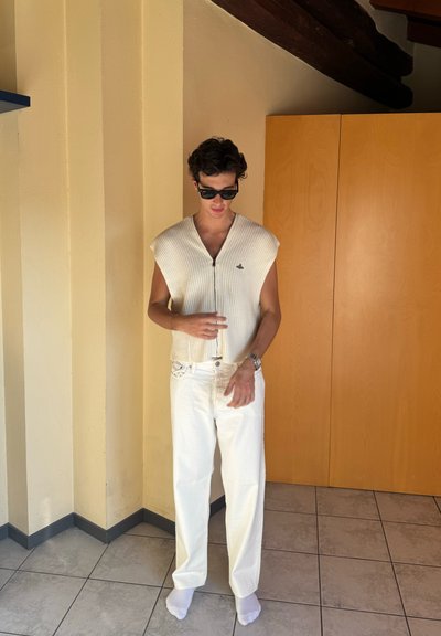 Top sin mangas de punto acanalado color marfil con cremallera en la parte delantera; combinado con pantalones anchos blancos. Los accesorios incluyen gafas de sol oscuras y un reloj plateado.