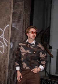 Pull camouflage gris et marron avec un col rond et un petit logo. Pantalon marron, lunettes rondes orange et chemise blanche à col visible.