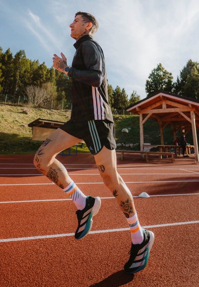 Atleta corriendo en una pista roja, vistiendo una chaqueta negra con cremallera y pantalones cortos, calcetines a rayas multicolores y zapatillas negras y verde azulado. Tatuajes visibles en las piernas.