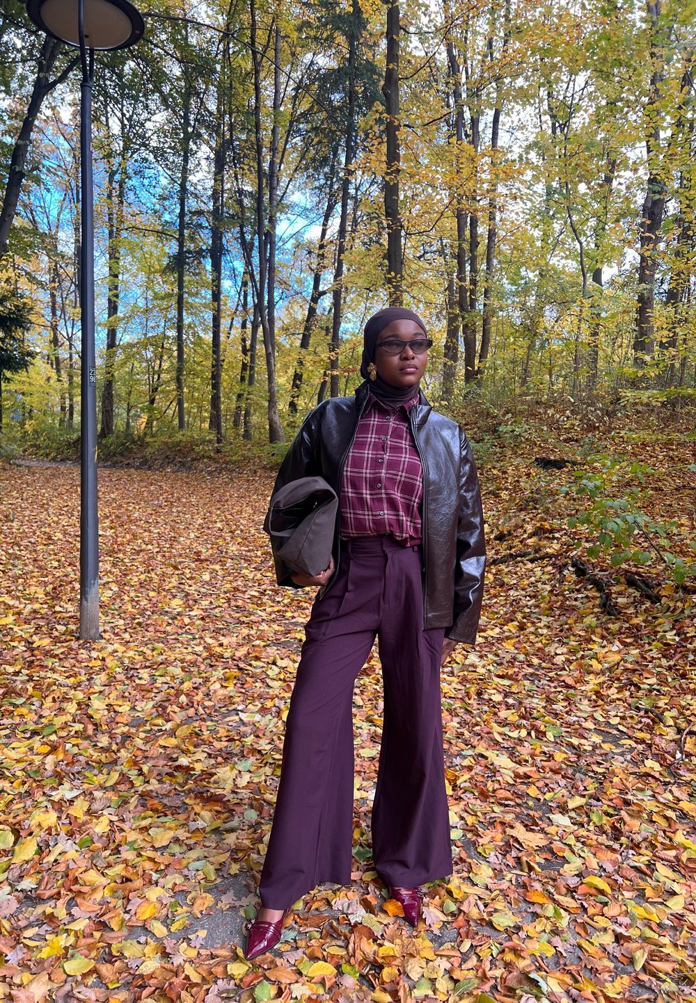 Modèle portant une veste en cuir marron, une chemise à carreaux rouges et un pantalon large bordeaux, accessoirisée avec des lunettes de soleil et de grandes boucles d'oreilles, dans un cadre forestier.