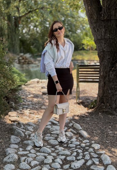 Camisa blanca de botones, falda negra con abertura frontal, cárdigan gris claro, zapatillas blancas y grises, y un pequeño bolso beige. Entorno informal al aire libre.