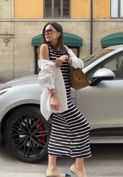 Mujer con gafas de sol, vestido de rayas, camisa blanca y bailarinas beige de pie junto a un coche plateado en una calle de la ciudad con edificios de piedra.