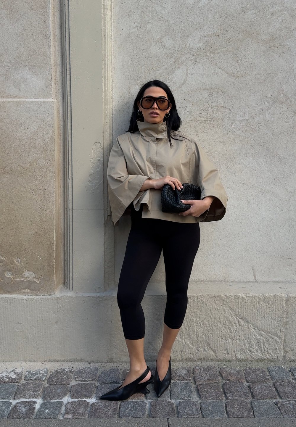 Beżowa krótka kurtka z szerokimi rękawami, zestawiona z czarnymi legginsami i butami na obcasie typu slingback. Trzyma czarną plecioną kopertówkę. Teksturowane tło ściany.