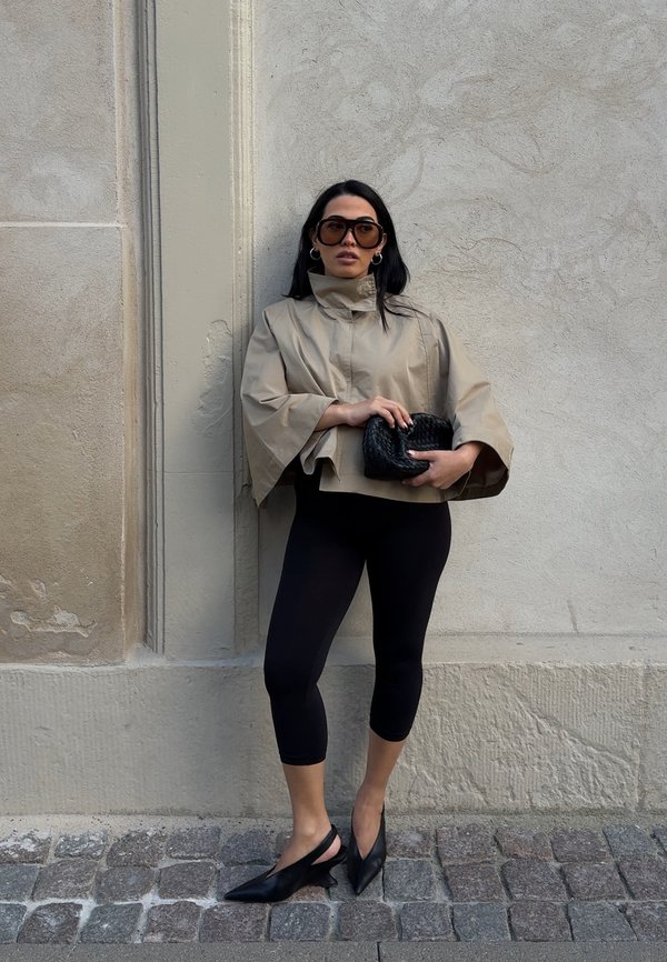 Beige cropped takki leveillä hihoilla, yhdistetty mustiin leggings-housuihin ja slingback-korkokenkiin. Pitelee mustaa kudottua laukku. Taitava seinä taustana.