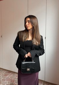 Sort faux læder håndtaske med krokodillestruktur, struktureret form og sølvspænde, siddende sammen med en lang lilla nederdel og sort blazer.