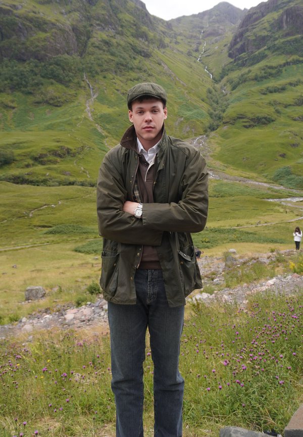 Junger Mann in grüner Jacke und Schiebermütze steht mit verschränkten Armen in einer grünen Gebirgslandschaft mit violetten Wildblumen und einem Wasserfall im Hintergrund.