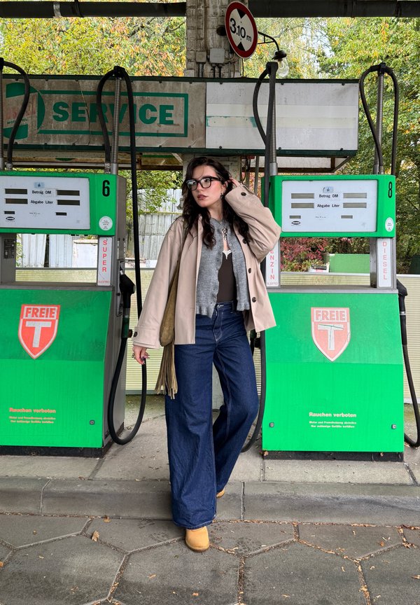 Een persoon staat naast groene benzinestations, gekleed in een beige jas over een grijze trui, wijdlopende blauwe jeans en tan schoenen.