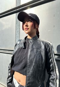 Μαύρο δερμάτινο κοντό μπουφάν με ασημί ρίγες, συνδυασμένο με ένα μαύρο crop top. Μια ασημί αλυσίδα κρέμεται από το φερμουάρ, κάτω από ένα μαύρο καπέλο.