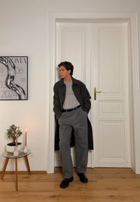 Manteau en laine gris sur un pull gris, pantalon large gris et chaussures noires. Sol en bois, petite plante et bougies à côté d'une table en verre.