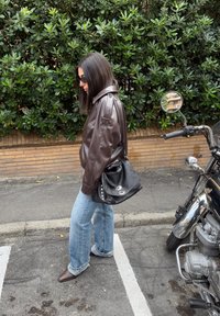 Barna bőr dzseki, túlméretezett világoskék farmernadrág és hegyes barna csizmák. Fekete kézitáska ezüst díszítéssel. Zöld növényzet és motoros háttér.