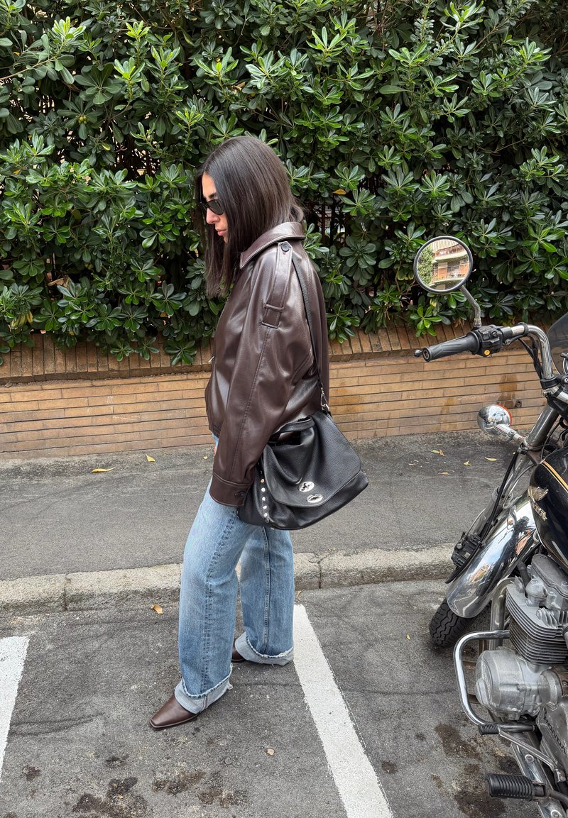 Casaco de couro castanho, jeans azul claro oversize e botas castanhas pontiagudas. Mala preta com detalhes prateados. Folhagem verde e fundo com motocicleta.