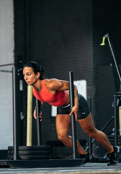 Atleta con una camiseta deportiva roja y pantalones cortos negros se apoya en una barra vertical, preparándose para un empuje, con discos de peso en primer plano.