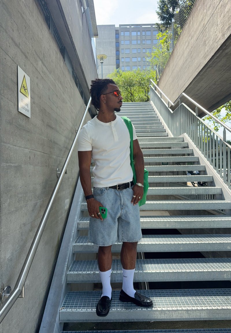 Uomo che indossa camicia bianca e pantaloncini di jeans, in piedi su scale metalliche, tiene un telefono verde e una borsa, guarda di lato all'aperto vicino a un muro di cemento.