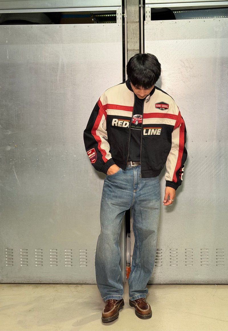 Jeune homme regardant vers le bas, portant une veste noire, blanche et rouge avec le texte "RED LINE", jean bleu et chaussures marron, debout contre une porte en métal.