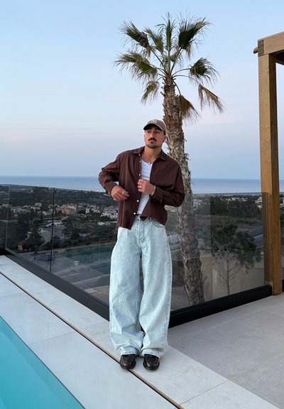 Camisa marrón de botón de corte relajado sobre una camiseta blanca, combinada con vaqueros de pierna ancha azul claro y zapatos negros en una terraza con una palmera.