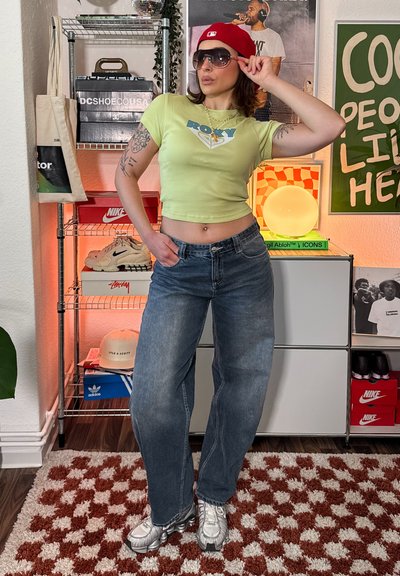 Mujer con gorra roja, gafas de sol, top corto verde y vaqueros anchos posa en una habitación con zapatillas deportivas, sombreros y pósteres enmarcados en estantes.