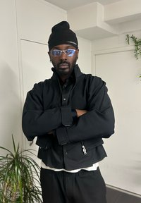 Veste noire raccourcie avec un col montant, poches zippées, superposée sur une chemise noire et un sous-vêtement blanc. Portée avec des lunettes de soleil et un bonnet.