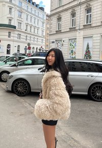 Ung kvinde i beige fluffy jakke og sort nederdel, der står på en byens gade med parkerede biler og juledekorationer i baggrunden.