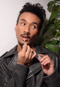 Veste en cuir noire, appliquant du gloss à lèvres avec un pinceau, accessoirisée avec des bagues et une boucle d'oreille, sur un fond simple avec de la verdure.