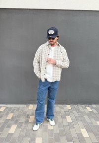 Man wearing a beige checked shirt, white T-shirt, blue jeans, trainers, sunglasses, and a navy Yankees cap standing on a tiled pavement.