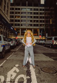 Isik, kes kannab halli dressipükse ja karvaga vooderdatud kapuutsiga jope, seisab linnatänava keskel kindlalt parkivate autode vahel.