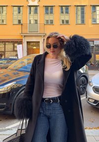 Abrigo de lana negro con mangas de piel texturizada sobre una blusa rosa clara, combinado con jeans de cintura alta azules y un cinturón negro. Unas gafas de sol redondas completan el look.