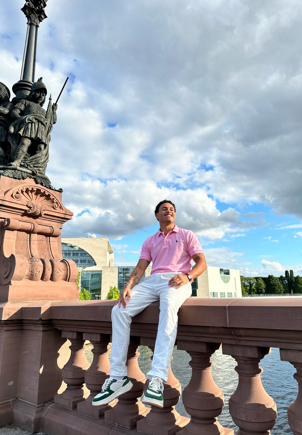 Man in a light pink polo shirt and white pants sits on a decorative stone railing near water. Green and white sneakers; cloudy sky backdrop.