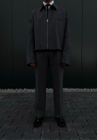 Σκούρο γκρι κοντό μπουφάν με μπροστινό φερμουάρ και μία τσέπη στο στήθος, συνδυασμένο με γκρι παντελόνι και μαύρα παπούτσια πάνω σε μοτίβο πεζοδρομίου.