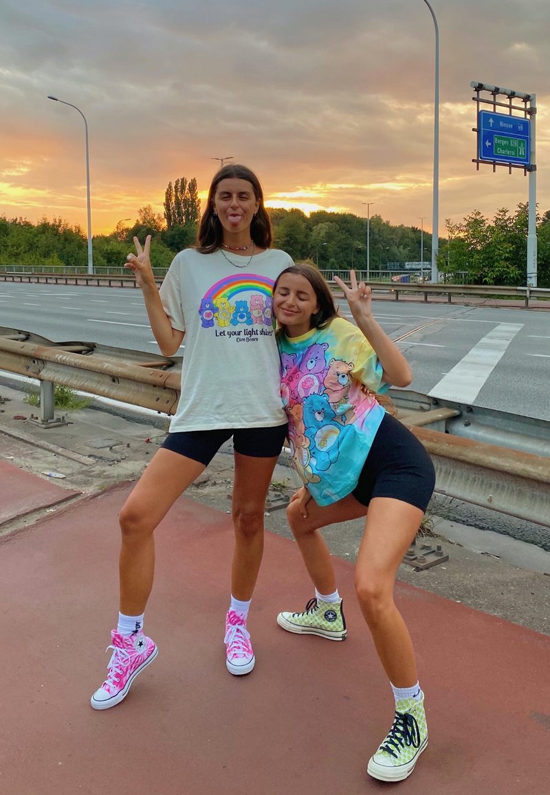 To personer poserer ved vejkanten iført grafiske t-shirts med farverige designs. Den ene har pink høj-top sneakers, mens den anden har grønne.