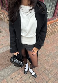 Schwarzer Wollmantel über einem weißen Pullover, graue Jeans-Shorts, schwarze Strumpfhosen und schwarze Mary Jane-Schuhe mit weißen Socken. Silberne Herz-Halskette und Handtasche.