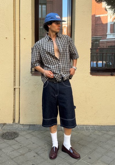 Camisa de manga corta a cuadros, pantalones cortos cargo de mezclilla oscura, calcetines blancos, mocasines marrones, gorra azul. Tejido texturizado, corte relajado, estilo casual.